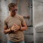 man in brown t-shirt sitting beside metal door and gray concrete wall