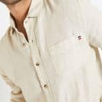 Un homme portant une chemise en lin beige froissé avec une étiquette indiquant la marque. La chemise est boutonnée et possède une poche à la poitrine, représentant un style casual-chic qui ne nécessite pas de repassage.